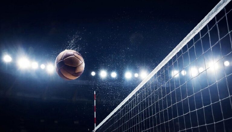 Волейбольный мяч пролетает над сеткой в луче прожектора на темном стадионе.