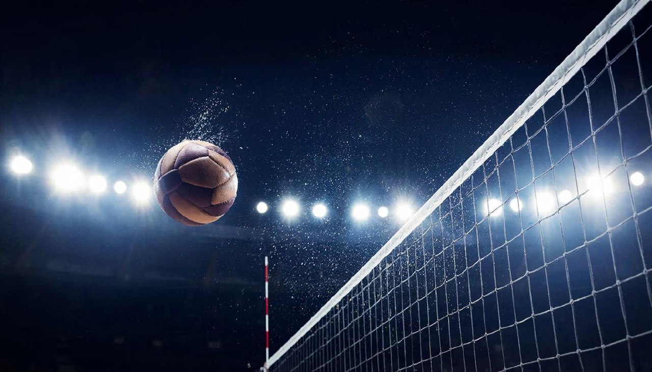 Волейбольный мяч пролетает над сеткой в луче прожектора на темном стадионе.